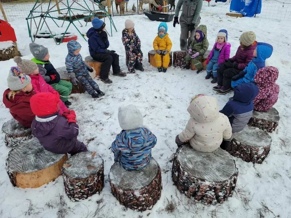 Children in snow gear sitting in a circle on stumps for morning circle